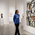 A woman in a blue hoodie examining a painting in a modern art gallery.